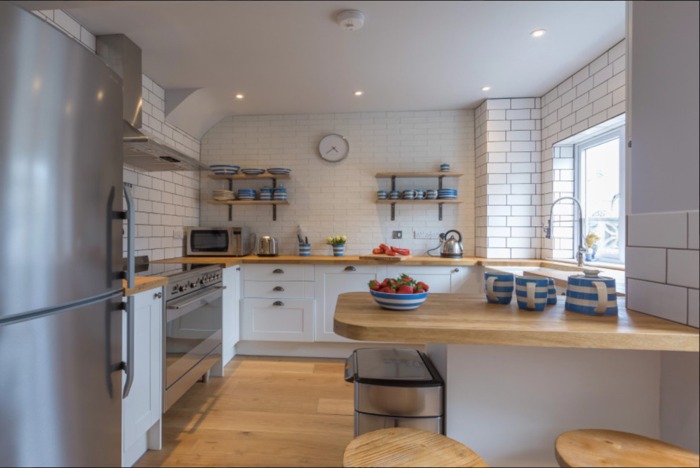 Mid-sized transitional kitchen in Cornwall with a farmhouse sink, shaker cabinets, white cabinets, wood benchtops, white splashback, subway tile splashback, stainless steel appliances, light hardwood floors, beige floor and beige benchtop.