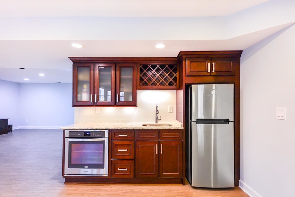 Photo of a mid-sized transitional single-wall wet bar in DC Metro with an undermount sink, raised-panel cabinets, dark wood cabinets, granite benchtops, vinyl floors, brown floor and beige benchtop.