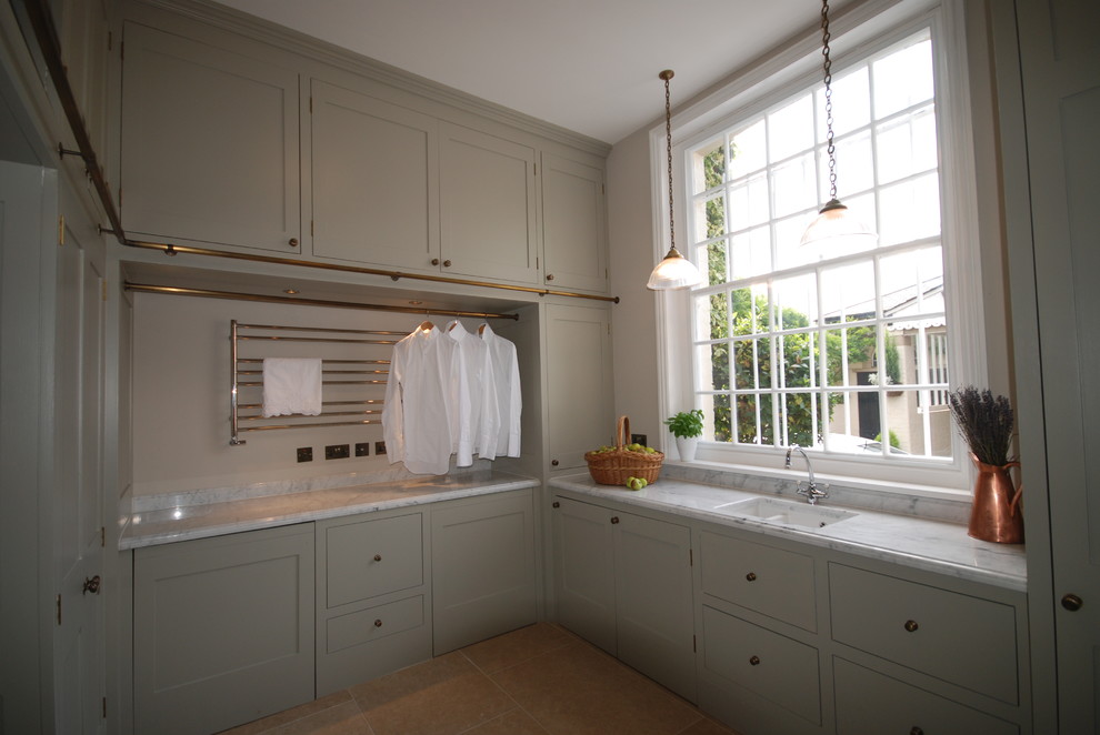 Photo of an expansive traditional u-shaped utility room in West Midlands with an undermount sink, shaker cabinets, grey cabinets, marble benchtops, grey walls, limestone floors, a concealed washer and dryer, beige floor and white benchtop.