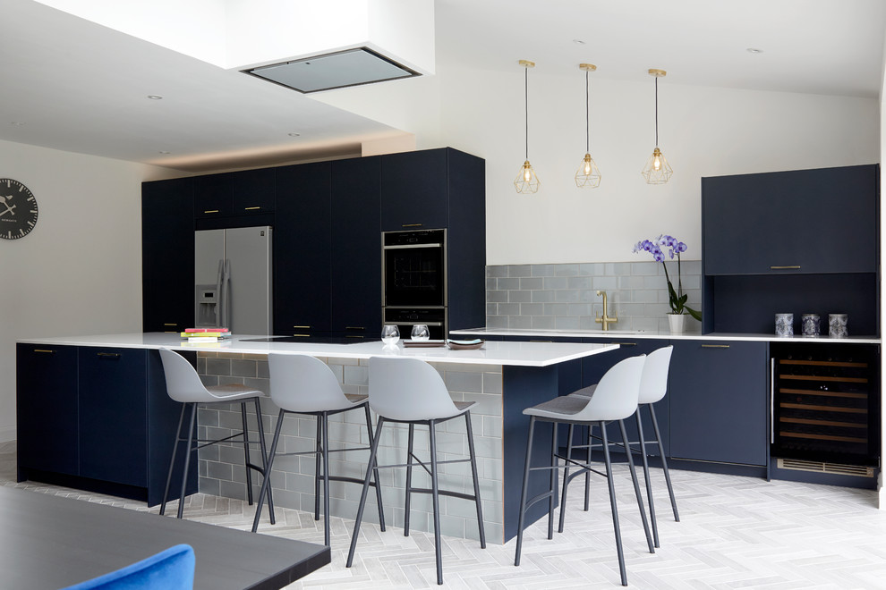 Photo of a large contemporary open plan kitchen in Kent with a drop-in sink, flat-panel cabinets, blue cabinets, solid surface benchtops, metallic splashback, glass tile splashback, black appliances, porcelain floors, with island, grey floor and white benchtop.