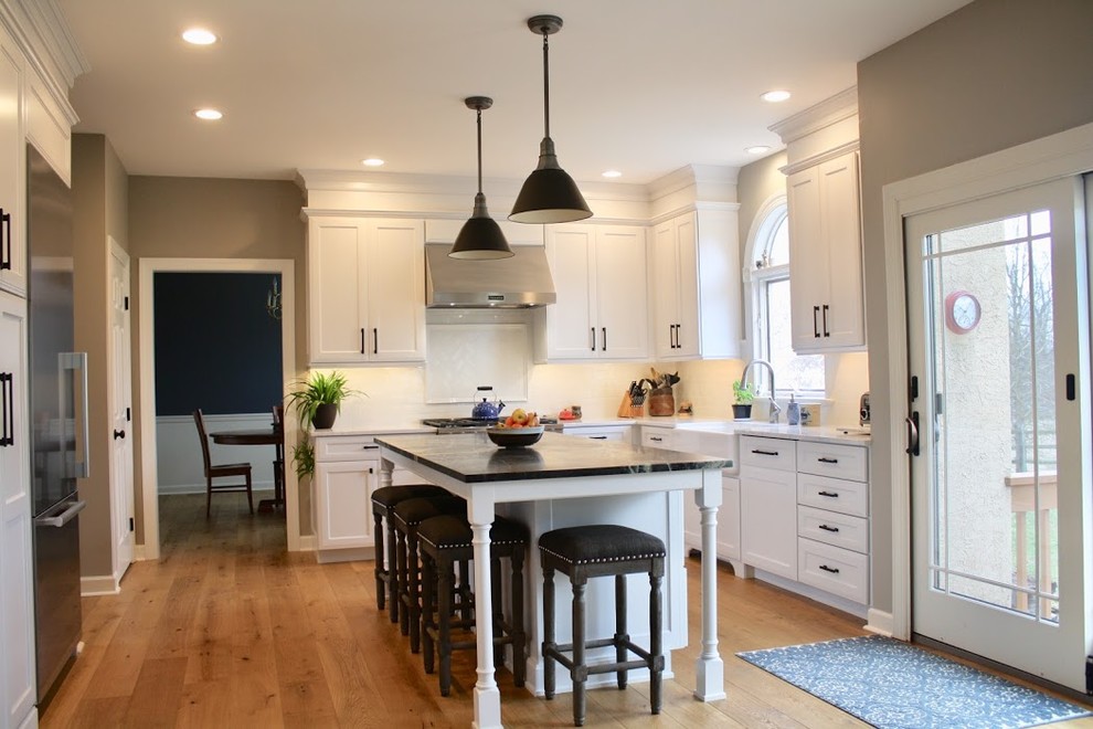 Photo of a mid-sized country l-shaped eat-in kitchen in Philadelphia with a farmhouse sink, recessed-panel cabinets, white cabinets, marble benchtops, white splashback, subway tile splashback, stainless steel appliances, medium hardwood floors, with island and brown floor.