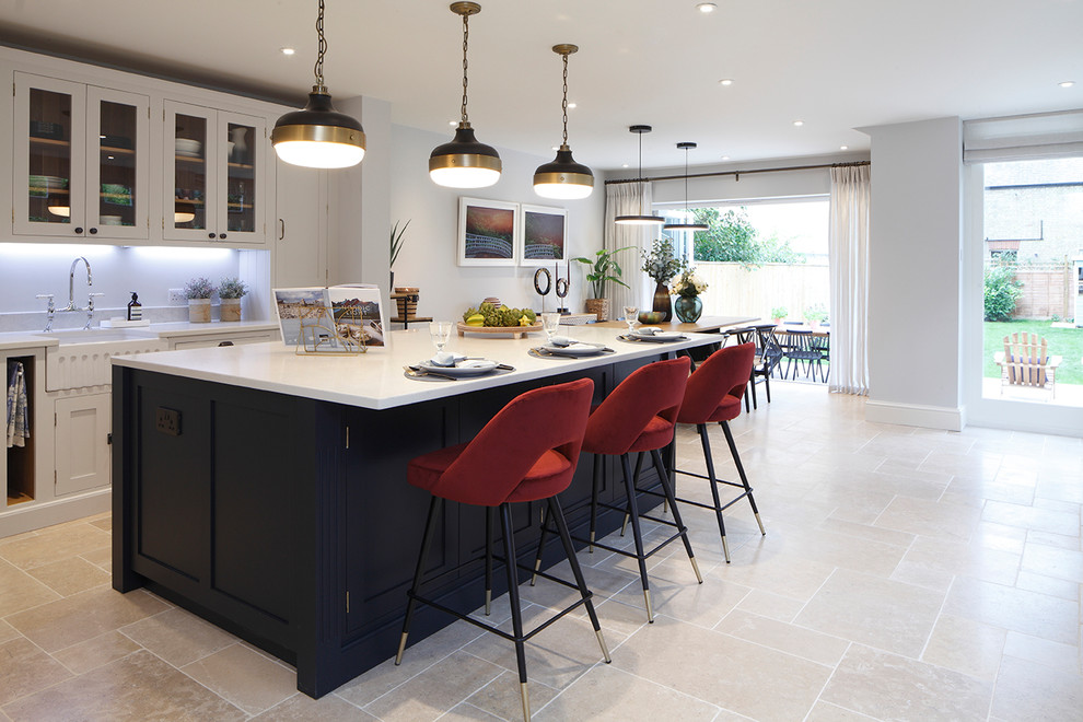 Large transitional l-shaped open plan kitchen in Oxfordshire with a farmhouse sink, shaker cabinets, white cabinets, solid surface benchtops, white splashback, black appliances, limestone floors, with island, beige floor and white benchtop.