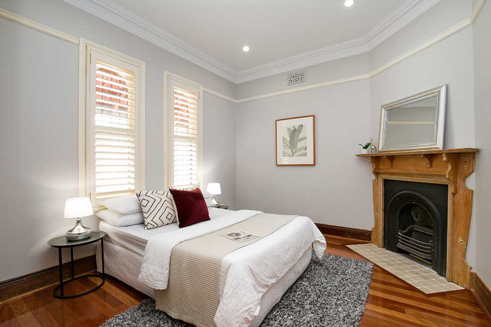This is an example of a transitional guest bedroom in Sydney with grey walls, medium hardwood floors, a corner fireplace, a wood fireplace surround and brown floor.