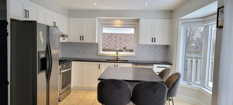 Transitional l-shaped eat-in kitchen photo with an undermount sink, recessed-panel cabinets, white cabinets, granite countertops, gray backsplash, porcelain backsplash, stainless steel appliances, an island and black countertops