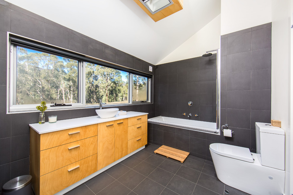 Photo of a mid-sized industrial kids bathroom in Melbourne with a drop-in sink, light wood cabinets, a two-piece toilet, porcelain tile, porcelain floors, furniture-like cabinets, a drop-in tub, a shower/bathtub combo, gray tile and grey walls.