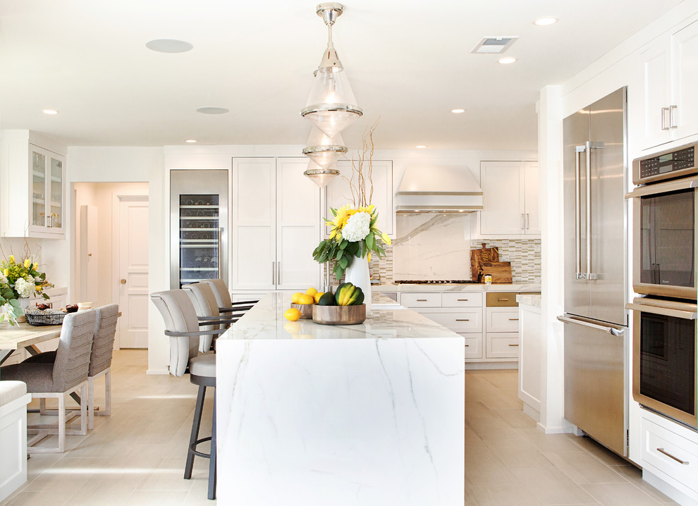 Mid-sized transitional l-shaped eat-in kitchen in Orange County with a farmhouse sink, shaker cabinets, white cabinets, quartz benchtops, white splashback, marble splashback, stainless steel appliances, porcelain floors, with island and beige floor.