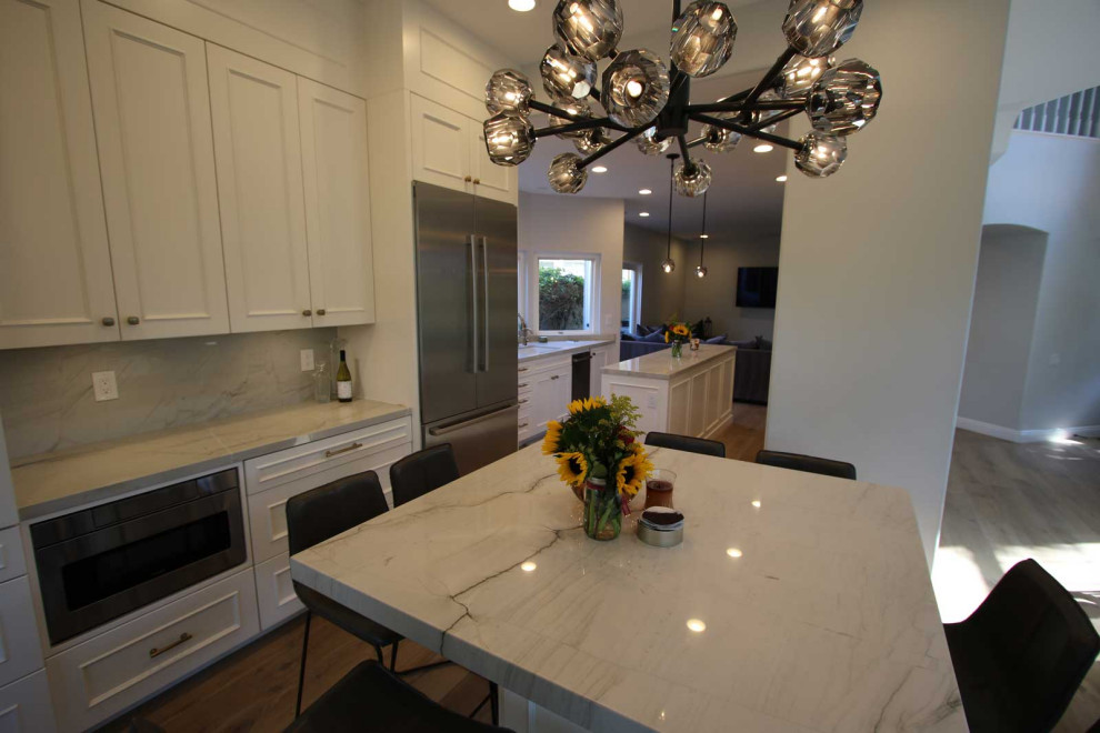 This is an example of a mid-sized transitional galley eat-in kitchen in Orange County with a farmhouse sink, shaker cabinets, white cabinets, granite benchtops, white splashback, granite splashback, stainless steel appliances, light hardwood floors, multiple islands, brown floor, white benchtop and wood.