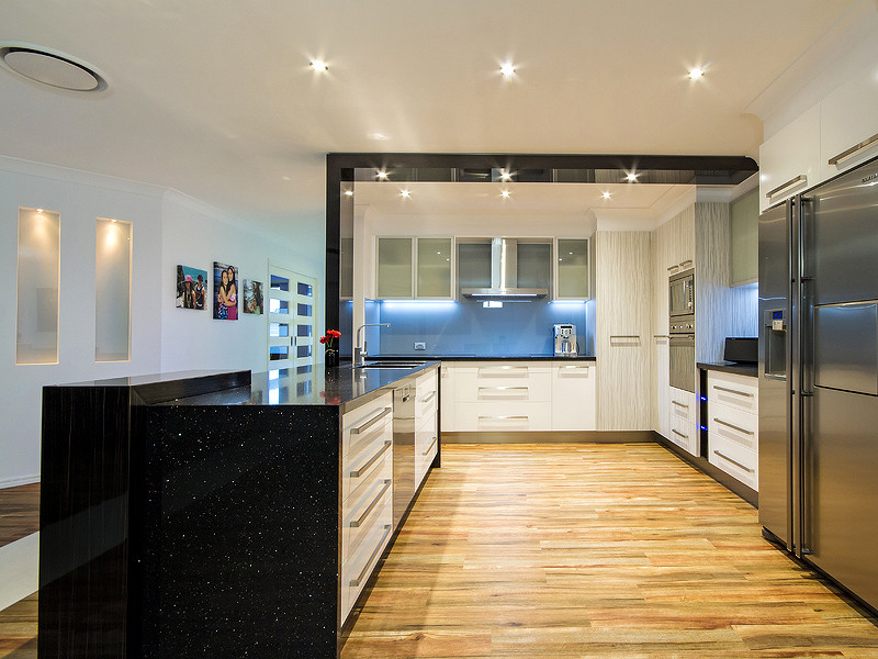 This is an example of a mid-sized contemporary u-shaped eat-in kitchen in Gold Coast - Tweed with a double-bowl sink, flat-panel cabinets, white cabinets, granite benchtops, blue splashback, glass sheet splashback, stainless steel appliances and light hardwood floors.