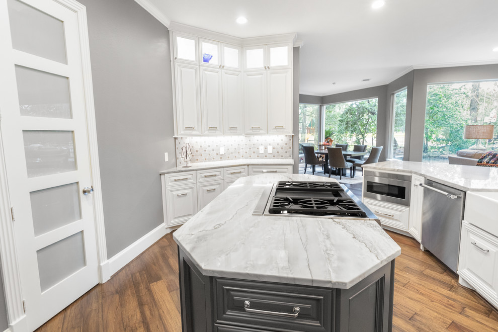 Photo of a large transitional u-shaped kitchen pantry in Houston with a farmhouse sink, raised-panel cabinets, grey cabinets, quartzite benchtops, grey splashback, limestone splashback, stainless steel appliances, dark hardwood floors, a peninsula, brown floor and grey benchtop.