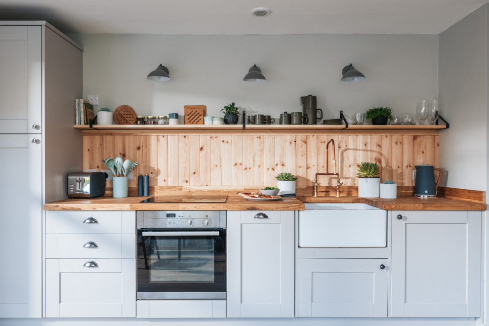 Inspiration for a medium sized rustic open plan kitchen in Hampshire with a belfast sink, recessed-panel cabinets, beige cabinets, wood worktops, wood splashback, stainless steel appliances and light hardwood flooring.