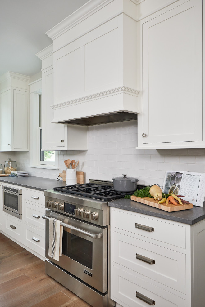 Photo of a mid-sized country l-shaped open plan kitchen in Grand Rapids with a farmhouse sink, recessed-panel cabinets, white cabinets, concrete benchtops, white splashback, subway tile splashback, stainless steel appliances, medium hardwood floors, with island and grey benchtop.
