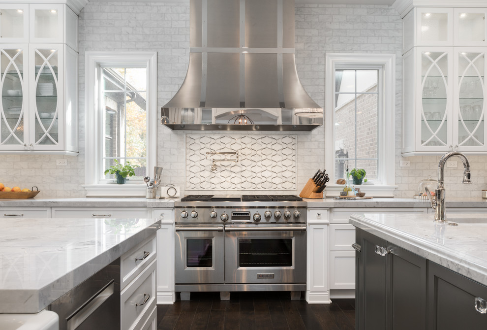 This is an example of a large transitional u-shaped separate kitchen in Chicago with a farmhouse sink, shaker cabinets, white cabinets, marble benchtops, white splashback, marble splashback, stainless steel appliances, dark hardwood floors, multiple islands and brown floor.