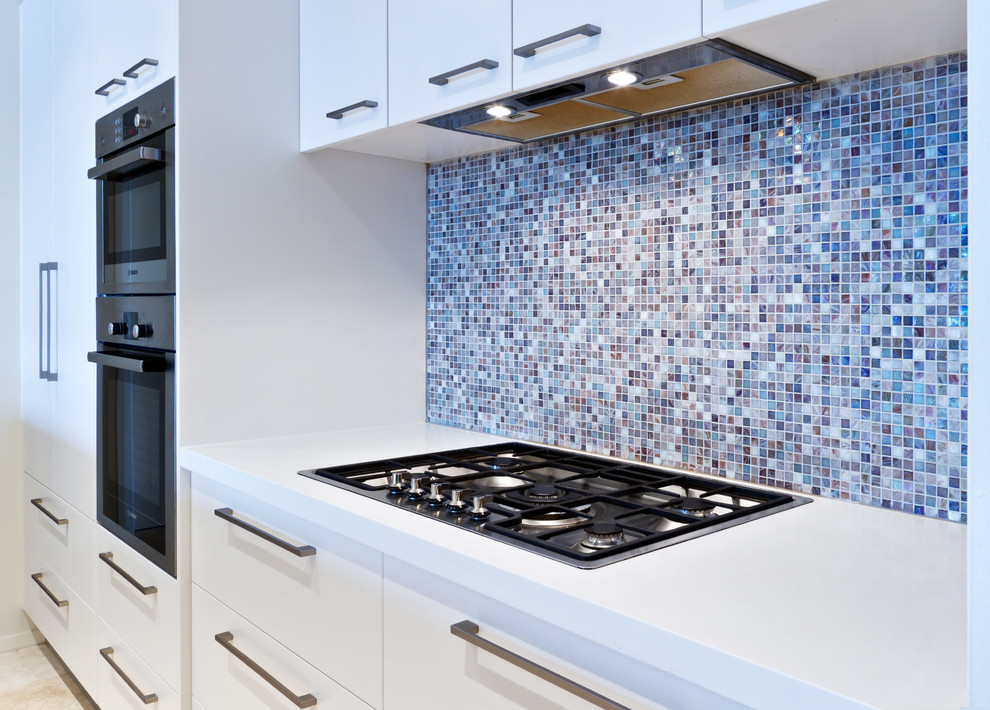 This is an example of a small modern galley kitchen in Sydney with flat-panel cabinets, white cabinets, quartz benchtops, multi-coloured splashback, mosaic tile splashback, stainless steel appliances, a double-bowl sink and travertine floors.