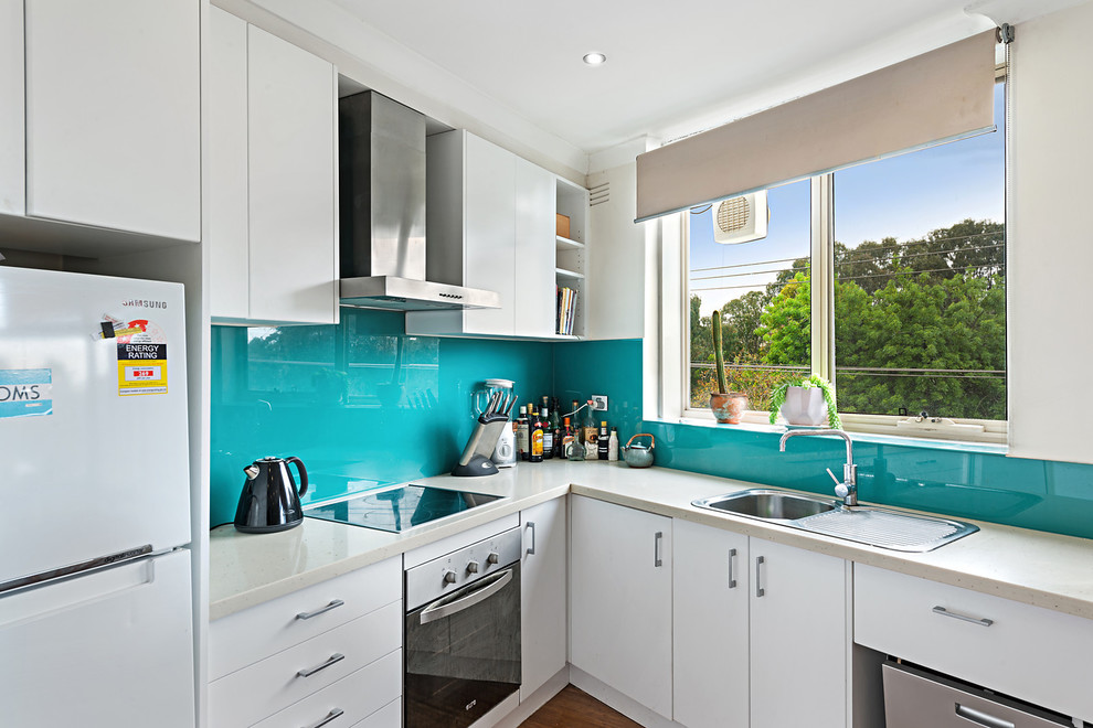 Small modern l-shaped open plan kitchen in Melbourne with a single-bowl sink, flat-panel cabinets, white cabinets, quartz benchtops, green splashback, glass sheet splashback, stainless steel appliances, bamboo floors and beige benchtop.
