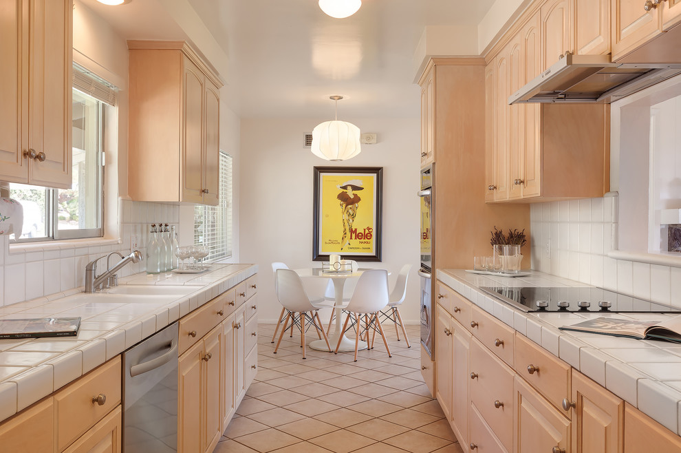 Photo of a small midcentury galley eat-in kitchen in Los Angeles with a double-bowl sink, raised-panel cabinets, light wood cabinets, tile benchtops, white splashback, porcelain splashback, stainless steel appliances, porcelain floors, beige floor and white benchtop.
