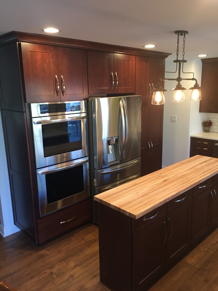Mid-sized transitional laminate floor and brown floor eat-in kitchen photo in Chicago with a farmhouse sink, recessed-panel cabinets, dark wood cabinets, quartz countertops, white backsplash, ceramic backsplash, stainless steel appliances and an island
