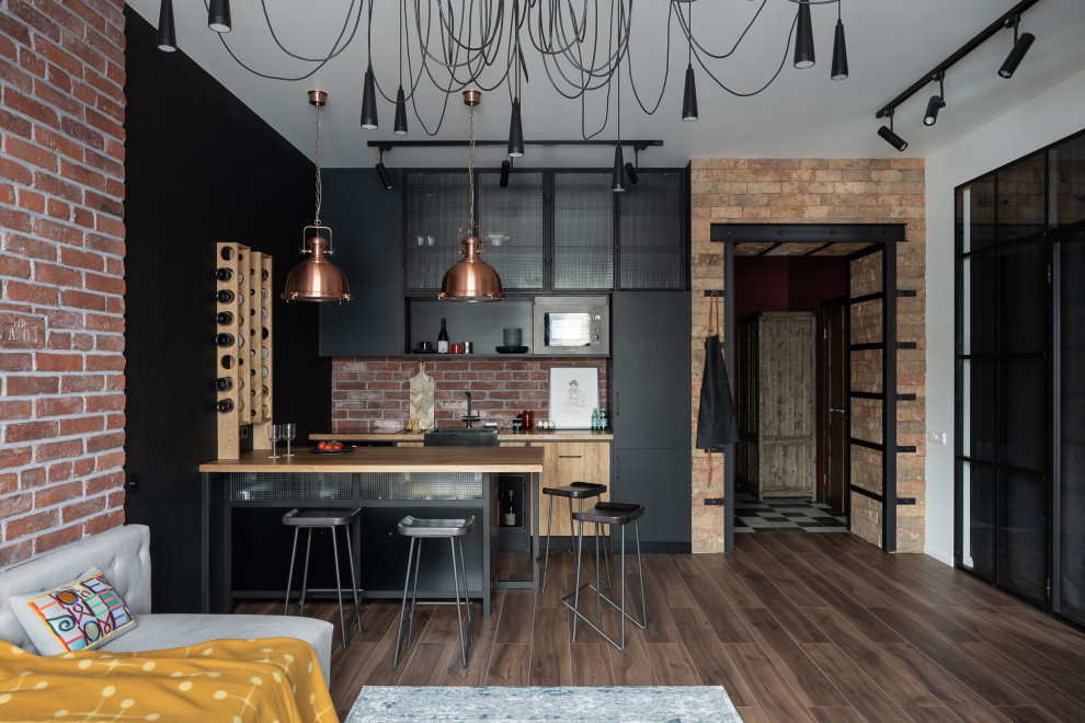 Inspiration for a mid-sized industrial u-shaped open plan kitchen in Saint Petersburg with black cabinets, wood benchtops, black appliances, brown floor, a farmhouse sink, flat-panel cabinets, red splashback, brick splashback, medium hardwood floors, a peninsula and beige benchtop.