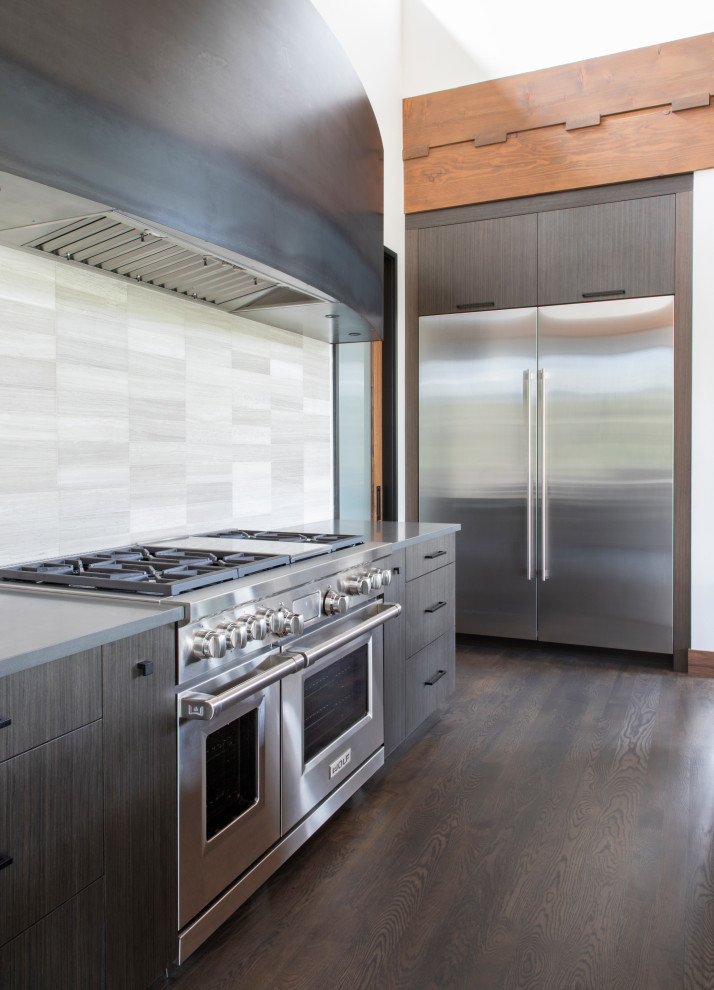 Example of a medium tone wood floor and vaulted ceiling kitchen design with an undermount sink, flat-panel cabinets, dark wood cabinets, quartz countertops, beige backsplash, porcelain backsplash, stainless steel appliances and an island