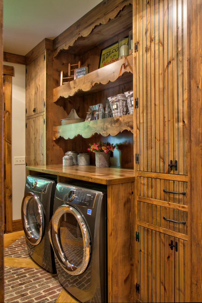 This is an example of a mid-sized country single-wall laundry room in Minneapolis with medium wood cabinets, wood benchtops, white walls, brick floors, a side-by-side washer and dryer, multi-coloured floor, brown benchtop and flat-panel cabinets.