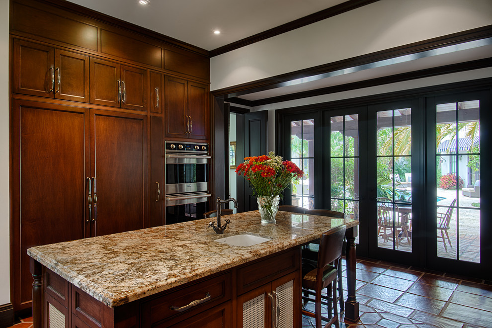 This is an example of a mid-sized mediterranean u-shaped separate kitchen in Miami with recessed-panel cabinets, dark wood cabinets, granite benchtops, panelled appliances, terra-cotta floors and with island.