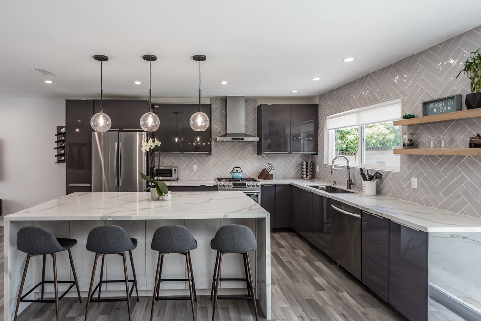 This is an example of an expansive contemporary l-shaped open plan kitchen in Los Angeles with an undermount sink, flat-panel cabinets, black cabinets, marble benchtops, white splashback, subway tile splashback, stainless steel appliances, vinyl floors, with island, multi-coloured floor and white benchtop.