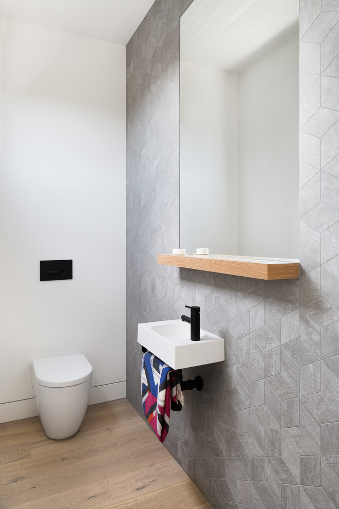 Photo of a contemporary powder room in Melbourne with a wall-mount toilet, gray tile, grey walls, light hardwood floors, a wall-mount sink and beige floor.