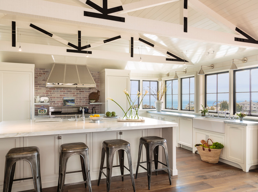 Design ideas for a beach style kitchen in Orange County with a farmhouse sink, shaker cabinets, white cabinets, brick splashback, stainless steel appliances, with island, grey benchtop, brown floor and medium hardwood floors.