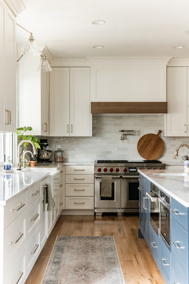 Inspiration for a medium sized classic u-shaped open plan kitchen in Salt Lake City with a belfast sink, recessed-panel cabinets, white cabinets, engineered stone countertops, white splashback, metro tiled splashback, stainless steel appliances, medium hardwood flooring, an island, multi-coloured floors and white worktops.
