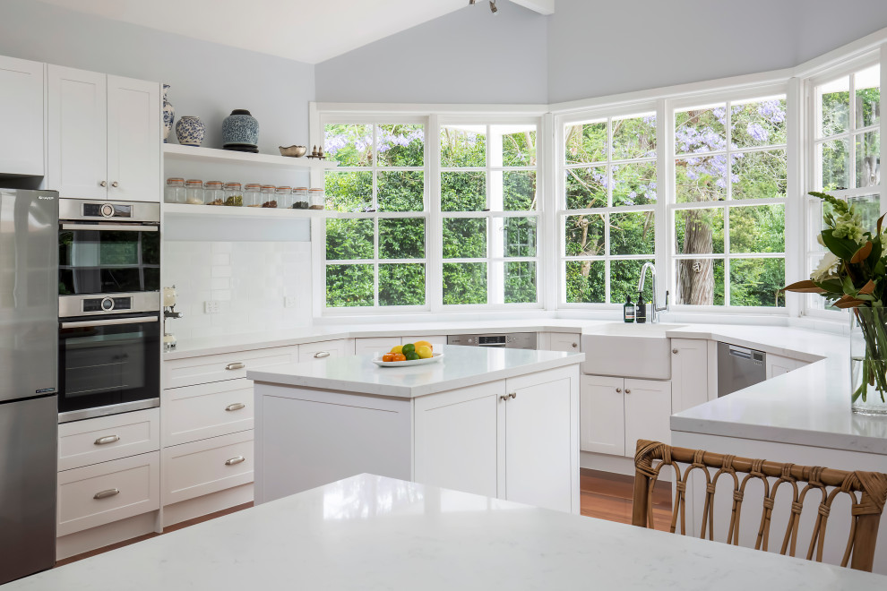 Inspiration for a large transitional u-shaped kitchen pantry in Sydney with a farmhouse sink, shaker cabinets, white cabinets, quartz benchtops, white splashback, subway tile splashback, stainless steel appliances, medium hardwood floors, multiple islands, brown floor, white benchtop and exposed beam.