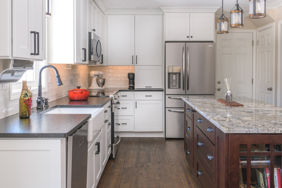 This is an example of a mid-sized transitional u-shaped eat-in kitchen in Boston with a farmhouse sink, recessed-panel cabinets, white cabinets, granite benchtops, white splashback, porcelain splashback, stainless steel appliances, with island, brown floor and black benchtop.