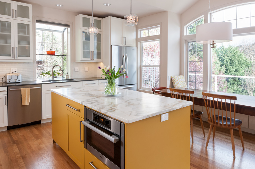 Inspiration for a mid-sized contemporary l-shaped open plan kitchen in Seattle with a double-bowl sink, shaker cabinets, yellow cabinets, marble benchtops, white splashback, glass tile splashback, stainless steel appliances, light hardwood floors and with island.