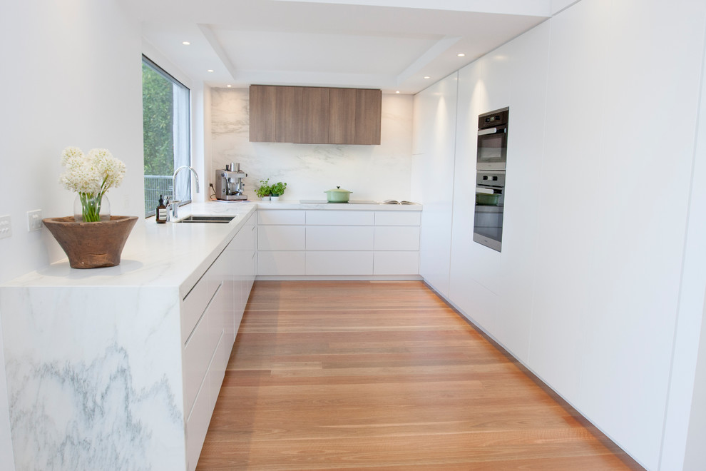 Photo of a contemporary u-shaped separate kitchen in Melbourne with an undermount sink, flat-panel cabinets, white cabinets, grey splashback, panelled appliances, medium hardwood floors, no island, brown floor and grey benchtop.