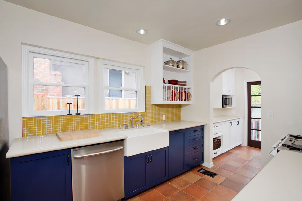 Design ideas for a mid-sized mediterranean galley separate kitchen in Los Angeles with a farmhouse sink, blue cabinets, yellow splashback, mosaic tile splashback, shaker cabinets, white appliances, terra-cotta floors and no island.