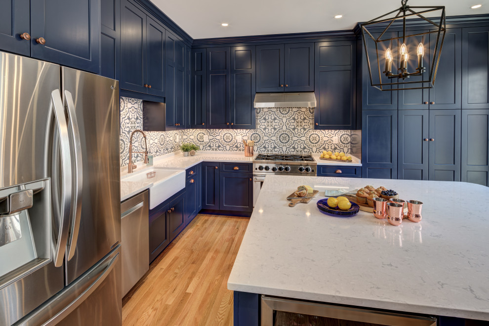 Example of a mid-sized transitional l-shaped medium tone wood floor and brown floor enclosed kitchen design in DC Metro with a farmhouse sink, shaker cabinets, blue cabinets, quartz countertops, multicolored backsplash, ceramic backsplash, stainless steel appliances, an island and white countertops