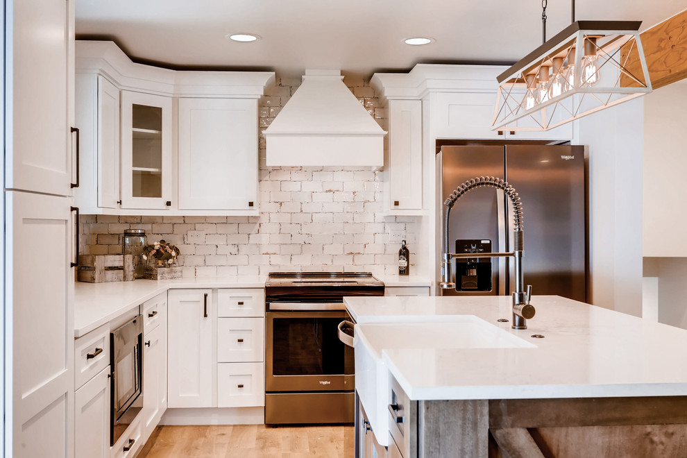 Photo of a large country l-shaped open plan kitchen in Denver with a farmhouse sink, shaker cabinets, white cabinets, quartz benchtops, white splashback, ceramic splashback, stainless steel appliances, laminate floors, with island, brown floor and white benchtop.