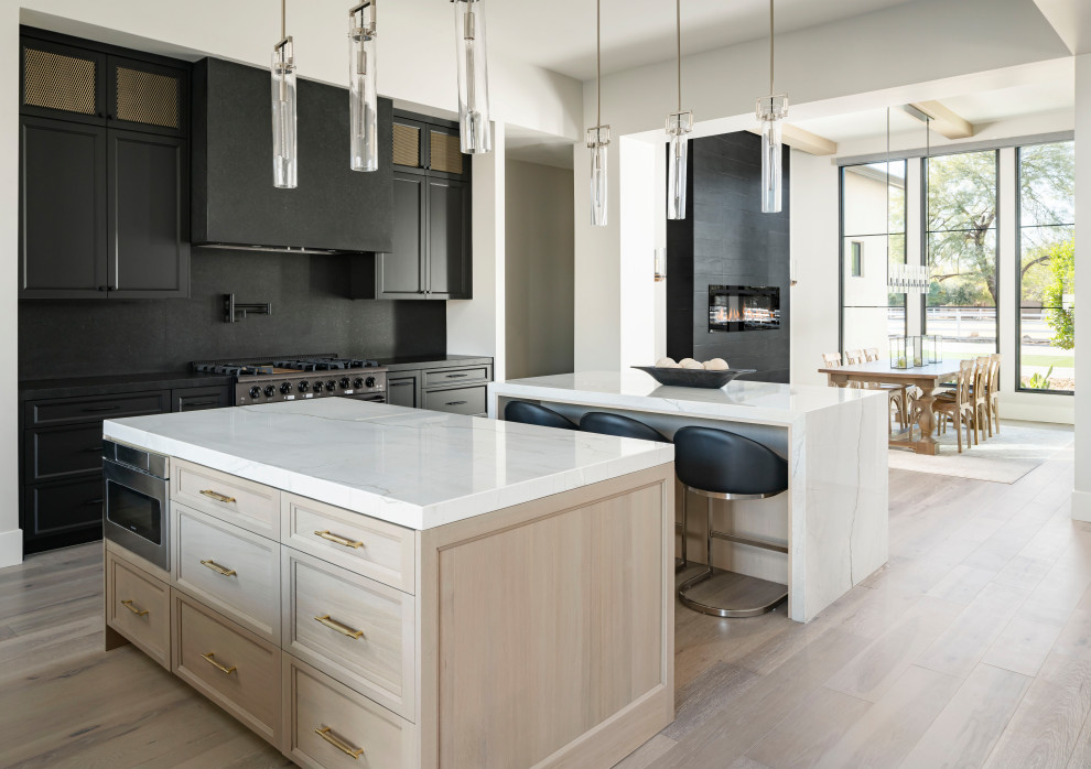 Inspiration for an expansive transitional l-shaped eat-in kitchen in Phoenix with an undermount sink, recessed-panel cabinets, medium wood cabinets, wood benchtops, black splashback, stone slab splashback, black appliances, light hardwood floors, multiple islands, grey floor and white benchtop.