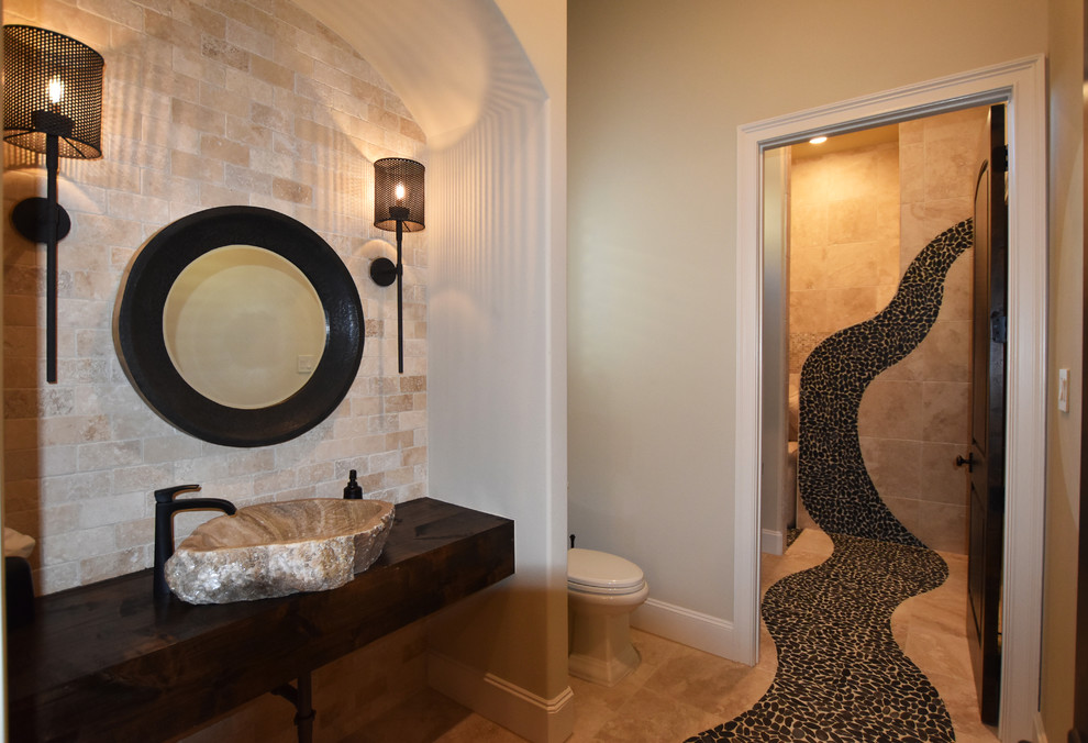 This is an example of a large mediterranean 3/4 bathroom in Houston with an alcove shower, a two-piece toilet, beige tile, stone tile, beige walls, pebble tile floors, a vessel sink and wood benchtops.
