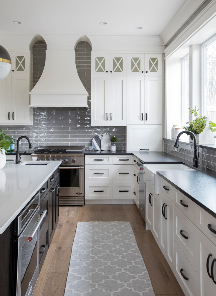 Country u-shaped eat-in kitchen in Other with a farmhouse sink, shaker cabinets, white cabinets, solid surface benchtops, grey splashback, brick splashback, stainless steel appliances, light hardwood floors, with island and brown floor.