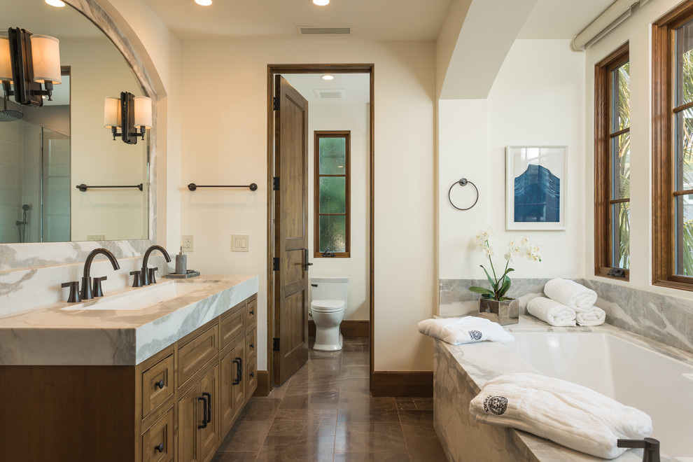 Mediterranean master bathroom in Orange County with recessed-panel cabinets, medium wood cabinets, an undermount tub, a one-piece toilet, beige walls, a trough sink, marble benchtops and brown floor.