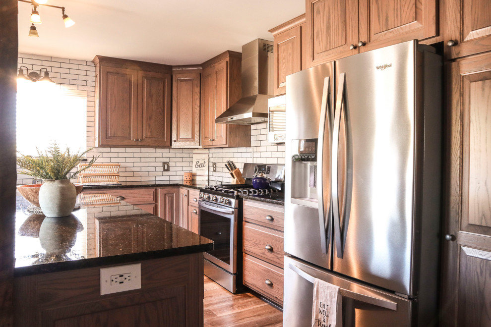 Mid-sized traditional u-shaped eat-in kitchen in Other with a farmhouse sink, raised-panel cabinets, brown cabinets, granite benchtops, white splashback, subway tile splashback, stainless steel appliances, vinyl floors, with island, brown floor and black benchtop.
