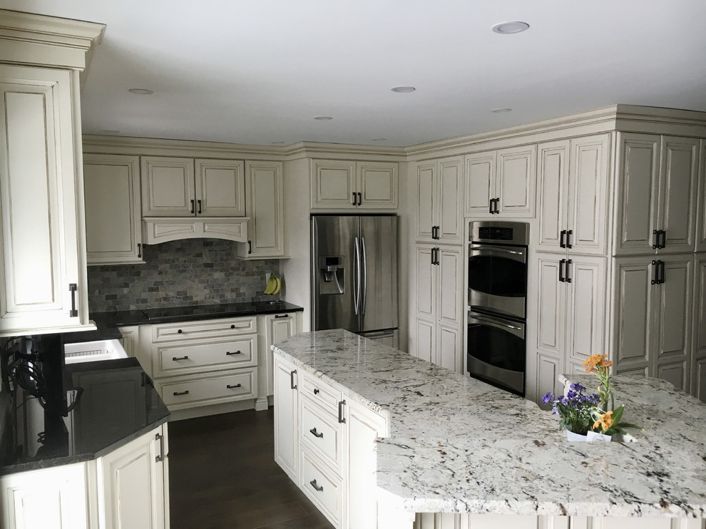 Photo of a large traditional u-shaped kitchen in Other with an undermount sink, raised-panel cabinets, white cabinets, granite benchtops, multi-coloured splashback, terra-cotta splashback, stainless steel appliances, dark hardwood floors, with island, brown floor and black benchtop.