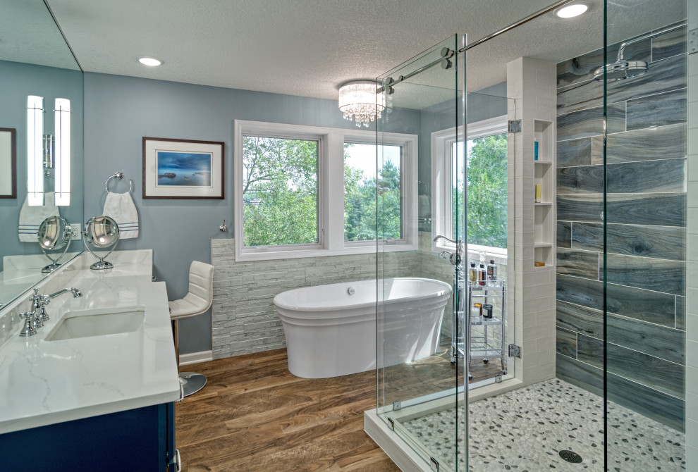 Photo of a large country master bathroom in Minneapolis with shaker cabinets, blue cabinets, a freestanding tub, a two-piece toilet, white tile, ceramic tile, grey walls, vinyl floors, an undermount sink, engineered quartz benchtops, brown floor, a sliding shower screen, white benchtops, a niche, a double vanity, a built-in vanity and decorative wall panelling.
