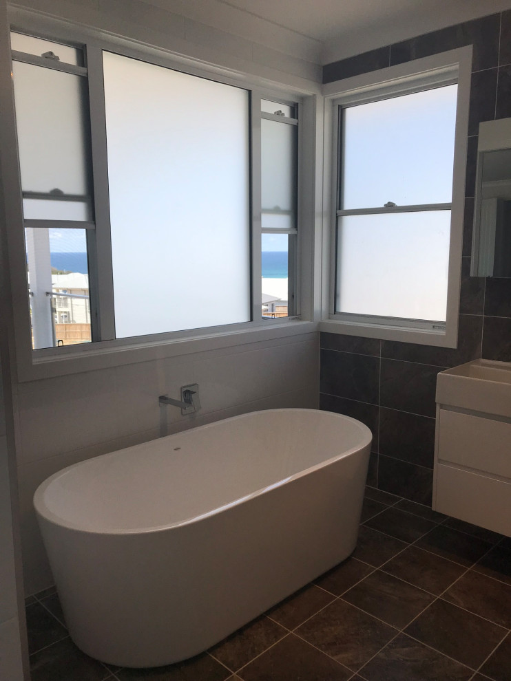 This is an example of a large beach style master bathroom in Central Coast with furniture-like cabinets, white cabinets, a freestanding tub, a double shower, a one-piece toilet, gray tile, porcelain tile, white walls, porcelain floors, a trough sink, solid surface benchtops, grey floor, an open shower and white benchtops.