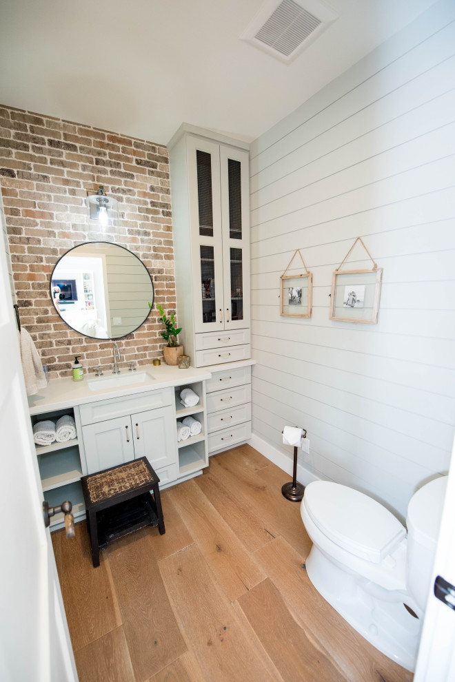 Mid-sized country powder room in San Diego with shaker cabinets, grey cabinets, a two-piece toilet, grey walls, medium hardwood floors, an undermount sink, quartzite benchtops, brown floor, white benchtops, a built-in vanity and brick walls.