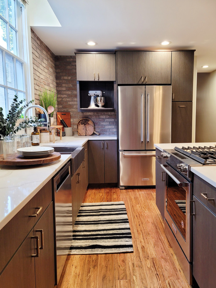 Photo of a mid-sized industrial l-shaped eat-in kitchen in New Orleans with a farmhouse sink, flat-panel cabinets, brown cabinets, quartzite benchtops, brown splashback, brick splashback, stainless steel appliances, bamboo floors, with island, brown floor and white benchtop.