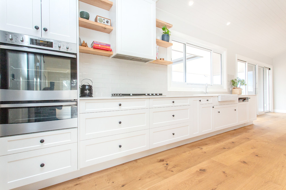 Design ideas for a mid-sized transitional galley open plan kitchen with a farmhouse sink, shaker cabinets, white cabinets, quartz benchtops, white splashback, subway tile splashback, stainless steel appliances, light hardwood floors, with island, brown floor and white benchtop.