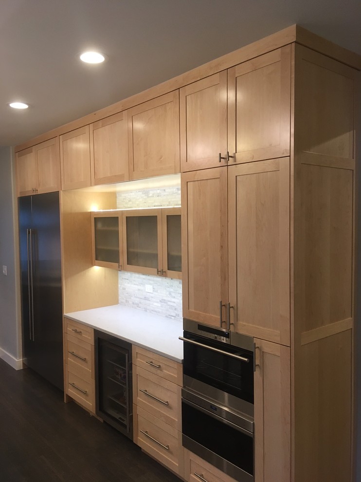 This is an example of a mid-sized transitional l-shaped kitchen in Chicago with a farmhouse sink, shaker cabinets, light wood cabinets, laminate benchtops, white splashback, glass tile splashback, white appliances, dark hardwood floors, with island and brown floor.