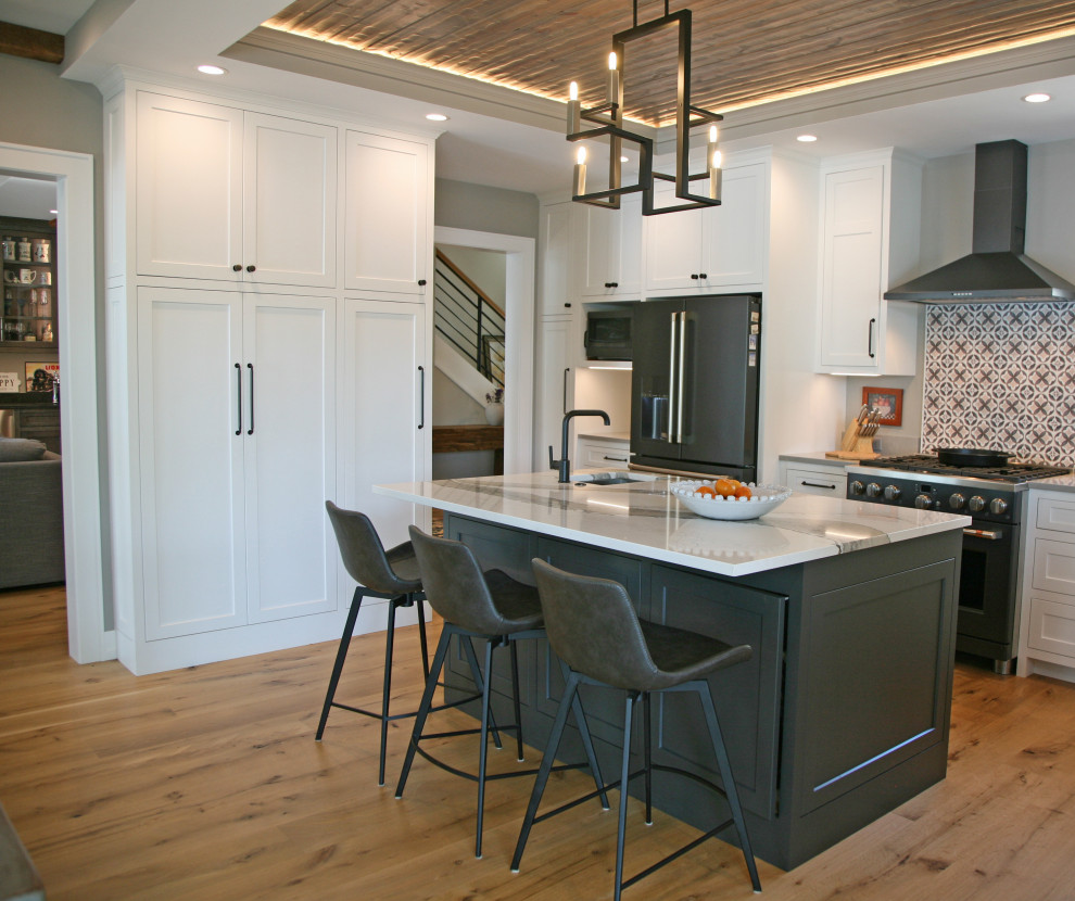 This is an example of a medium sized rural l-shaped kitchen/diner in Milwaukee with a belfast sink, shaker cabinets, black cabinets, engineered stone countertops, multi-coloured splashback, mosaic tiled splashback, black appliances, medium hardwood flooring, an island, brown floors, white worktops and a wood ceiling.