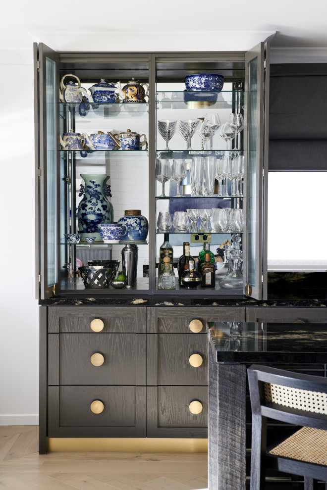 Photo of a mid-sized eclectic galley home bar in Other with an undermount sink, glass-front cabinets, grey cabinets, granite benchtops, black splashback, granite splashback, medium hardwood floors, grey floor and black benchtop.