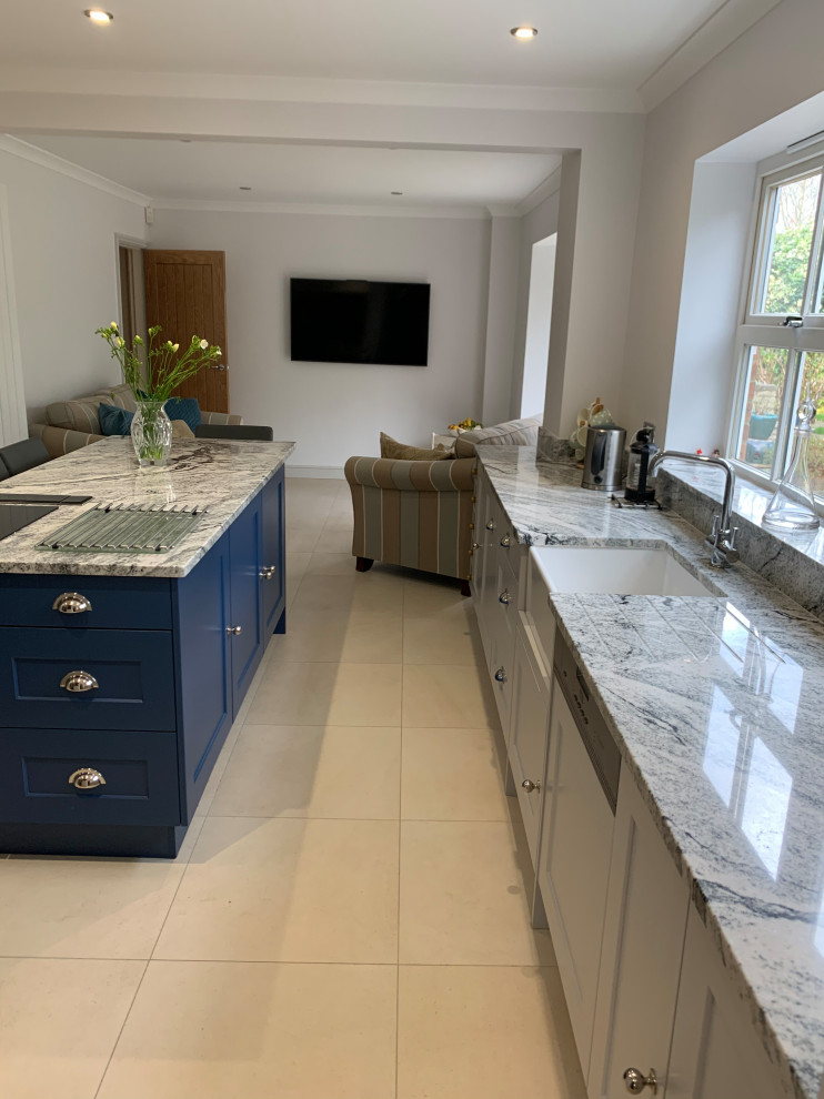 This is an example of a large l-shaped open plan kitchen in Sussex with a farmhouse sink, shaker cabinets, grey cabinets, granite benchtops, grey splashback, granite splashback, black appliances, porcelain floors, with island, beige floor and grey benchtop.